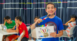 preteen kids at computers and one boy in the front facing camera holding a movie clapboard and smiling